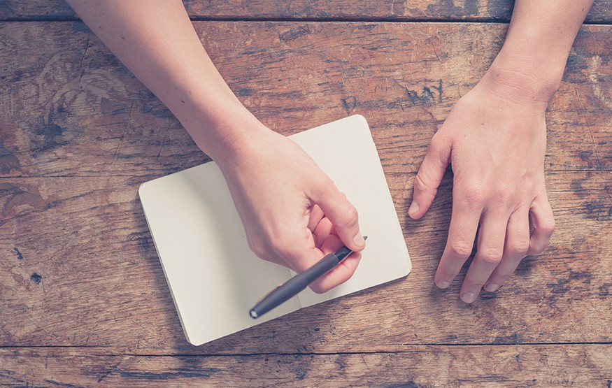 Close up on the hands of a young woman as she is writing in a small notepad at a wooden table